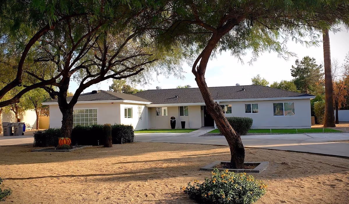 Exterior view of a single-story white building with a dark roof, surrounded by trees and landscaping. The foreground shows a dry, sandy area with some bushes and a curved tree trunk framing the building.