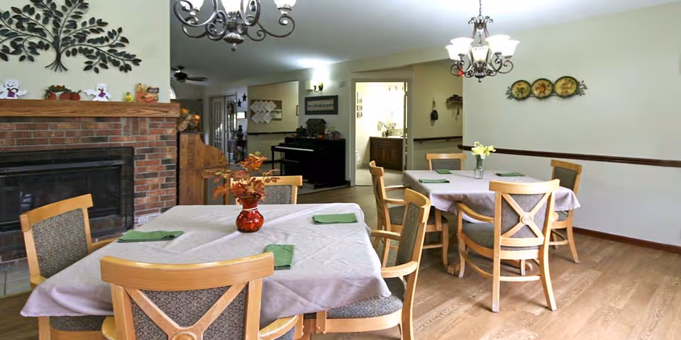 Communal dining room with several round tables and wooden chairs set with tablecloths and centerpieces, a brick fireplace and wall decor.