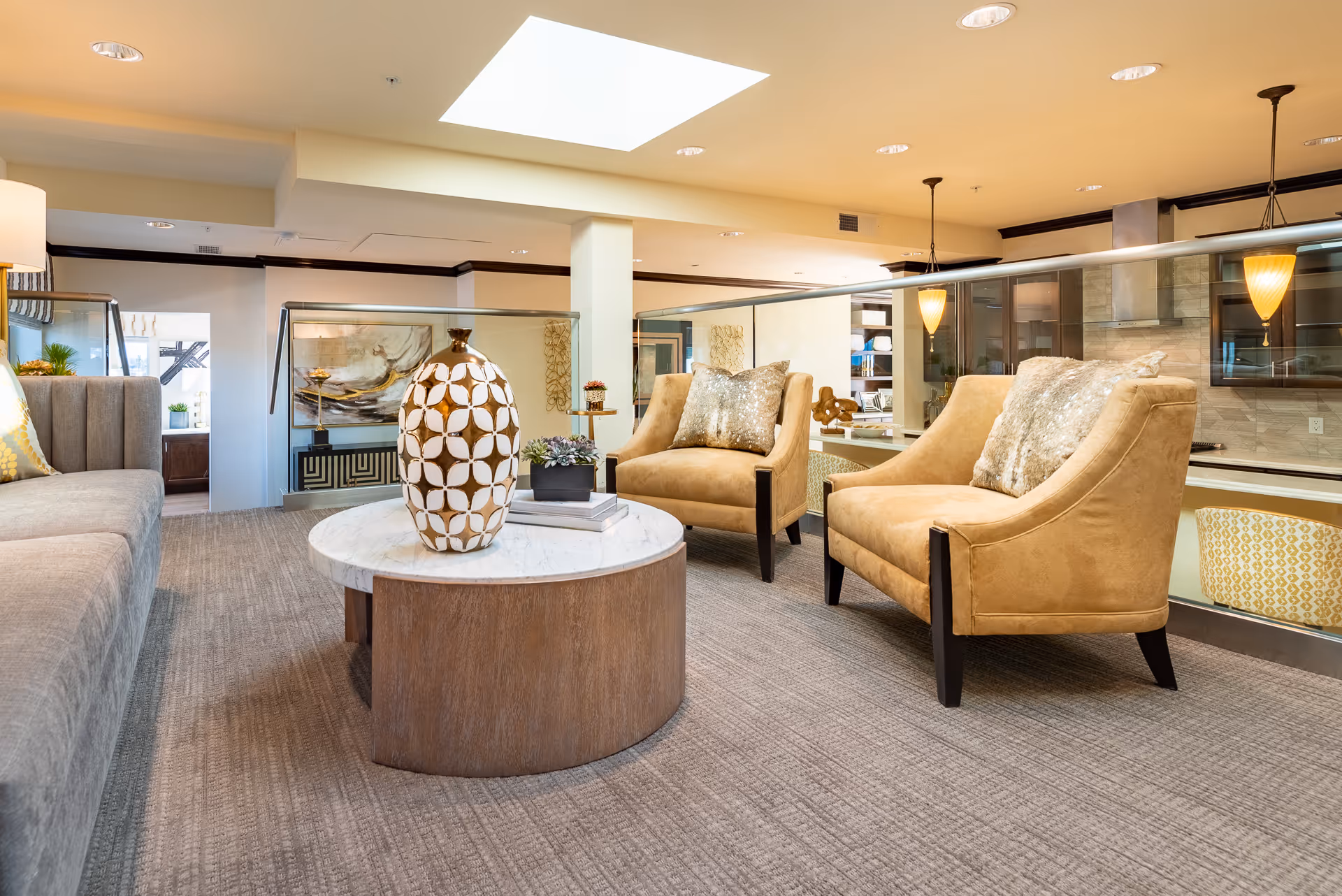 A cozy senior living common area with a gray sofa on the left, two tan armchairs with decorative pillows on the right, and a round wooden coffee table with a white marble top in the center. The table holds a large decorative vase and a small plant. The room has beige walls, carpeted floor, recessed lighting, and a skylight above. A glass railing separates this area from a kitchen space visible in the background.