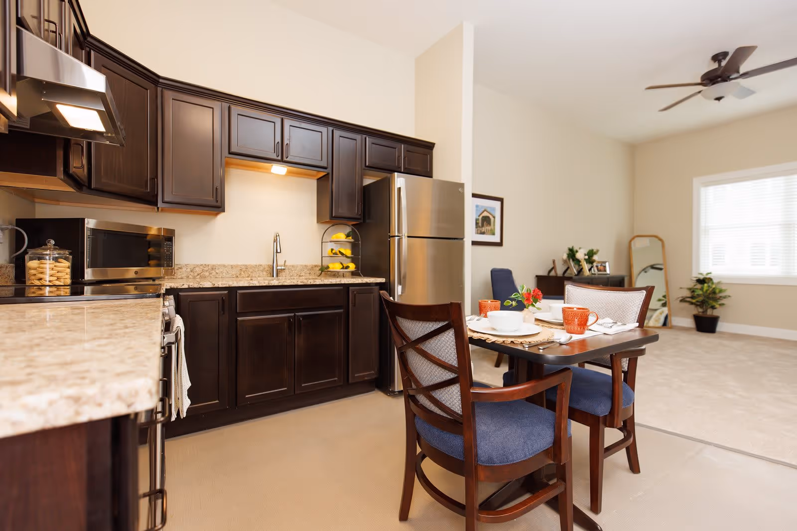 Open-plan kitchen with dark wood cabinets, stainless refrigerator, and a small dining table set for two opening into a living area.