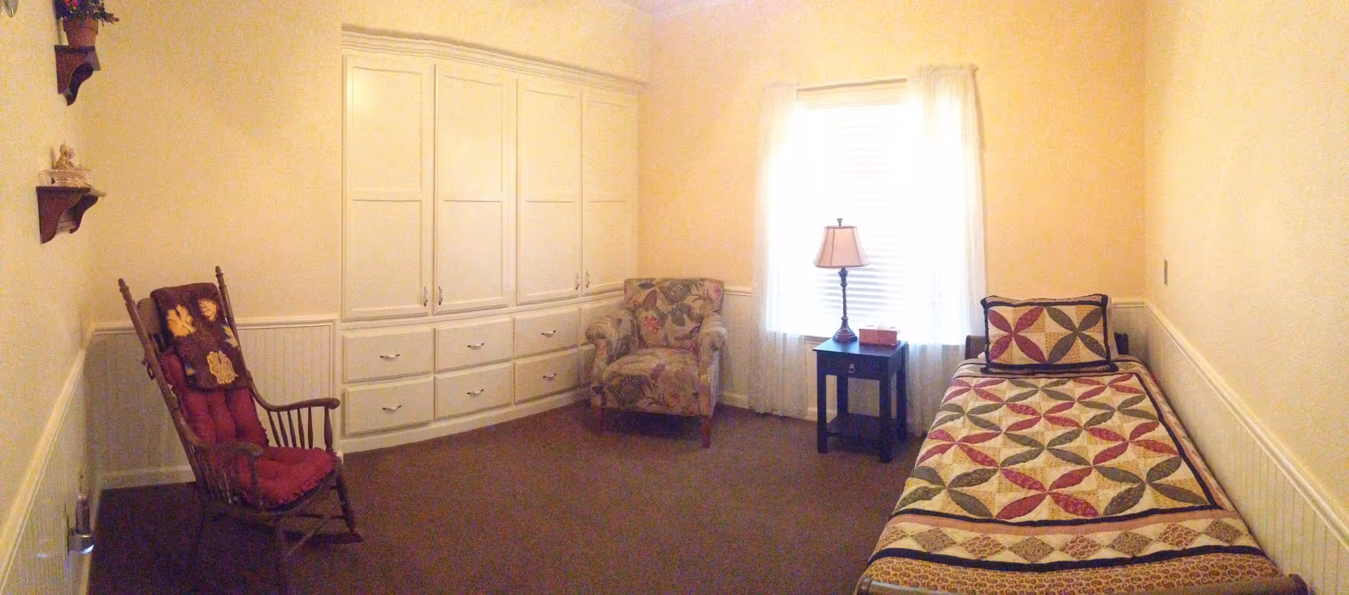 A small, cozy bedroom with a single bed covered in a patterned quilt, a wooden rocking chair with a cushion and throw, a floral upholstered armchair, a small dark wooden side table with a lamp and tissue box, and built-in white cabinets along one wall. The room has beige walls and carpet, with a window covered by sheer white curtains.