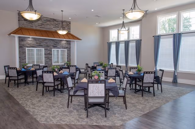 A bright dining room with multiple tables covered in dark tablecloths, each set with napkins and small plants. The room features large windows with blue curtains, pendant lights hanging from the ceiling, and a decorative stone wall with a copper-colored canopy above two windows.