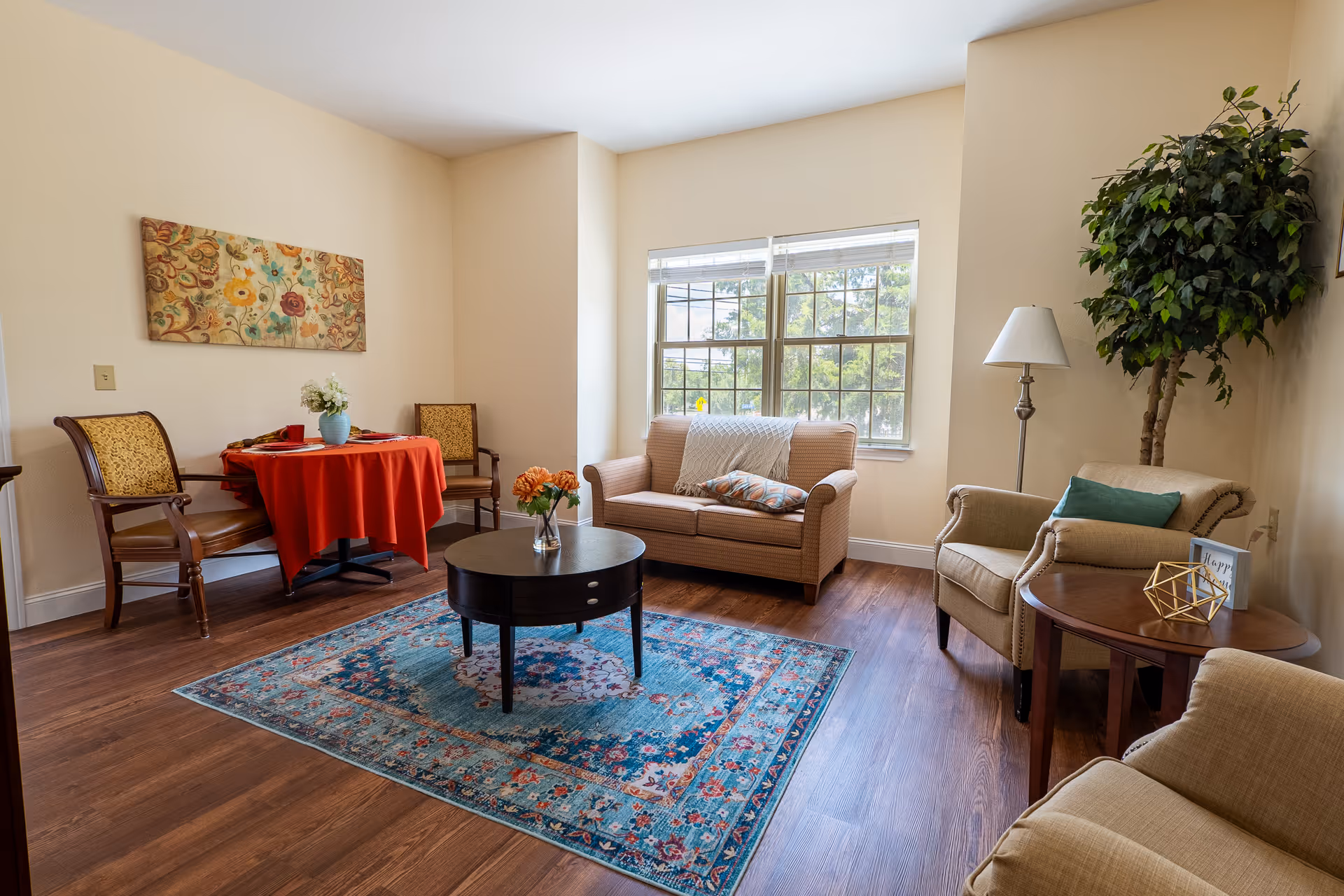 A cozy living room with a beige loveseat and two beige armchairs arranged around a round wooden coffee table on a blue patterned rug. There is a small round table with a red tablecloth and two wooden chairs with yellow patterned cushions against the wall. A large window lets in natural light, and a tall green plant stands in the corner next to a floor lamp. A floral painting hangs on the wall above the table.