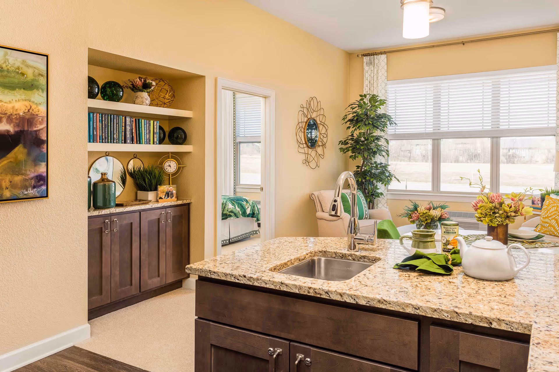 Bright and cozy interior space featuring a granite kitchen island with a sink and faucet, decorated with a white teapot, green napkins, and a small bouquet of flowers. In the background, there is a comfortable seating area with a beige armchair, green cushions, a tall potted plant, and large windows with white blinds letting in natural light. A built-in bookshelf with books, decorative items, and a clock is visible on the left, along with a doorway leading to a bedroom with a green bedspread.