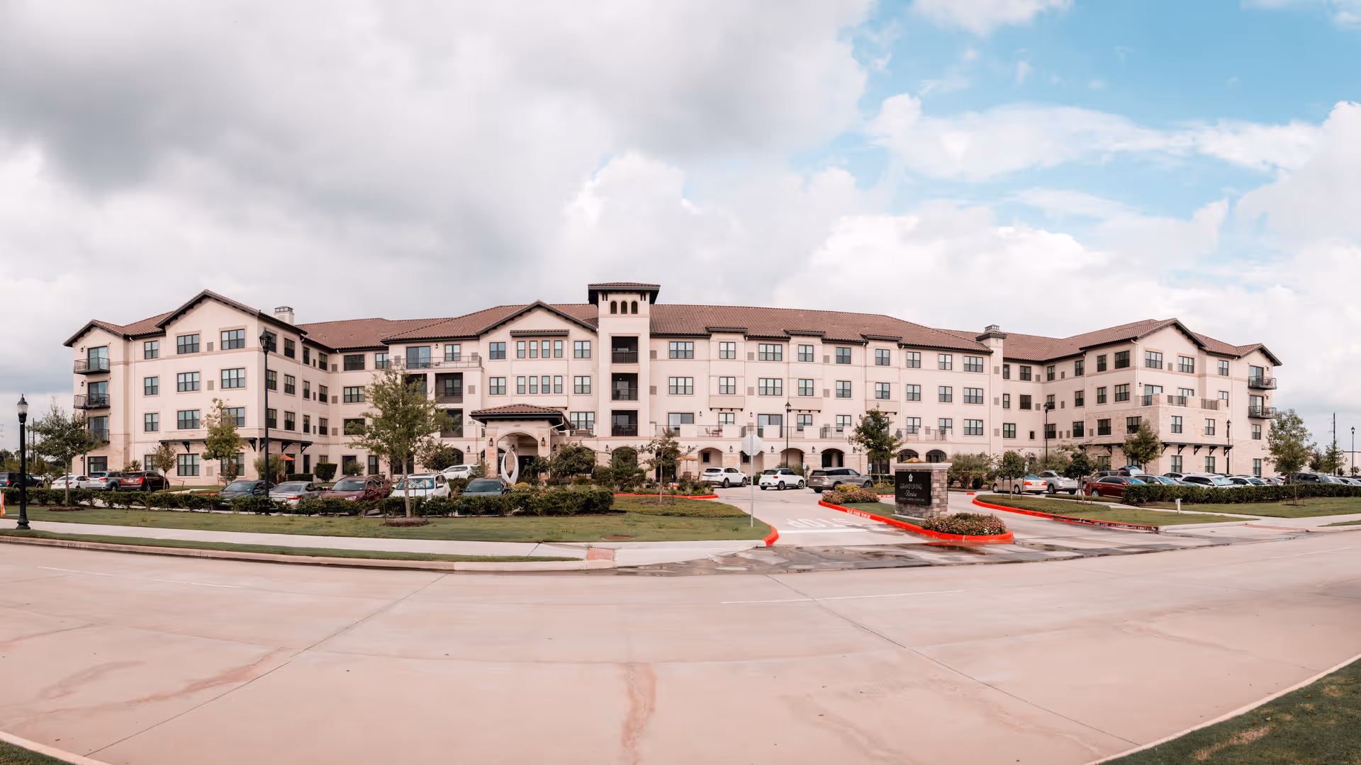 Wide exterior view of a large, multi-story senior living facility building with beige walls and a red-tiled roof, surrounded by landscaped greenery and a parking lot with several cars. The sky is partly cloudy with patches of blue.
