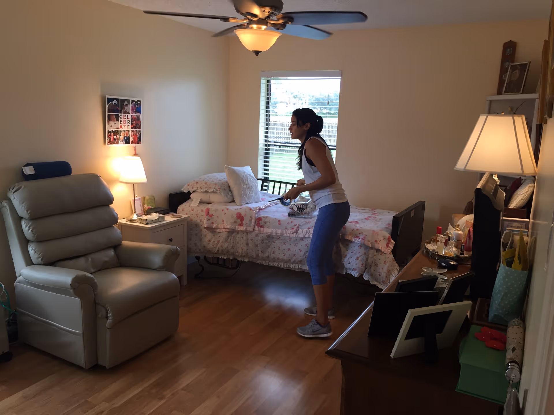 A woman is making a bed in a cozy bedroom with wooden flooring. The room features a beige recliner chair, a bedside table with a lamp, a window with blinds, and a dresser with a lamp and various personal items. The bed has floral bedding and several pillows.