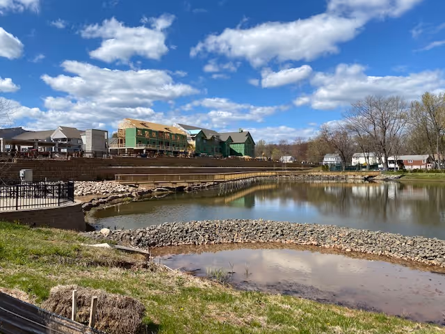 View of a partially constructed building complex next to a calm pond with a stone embankment under a blue sky with scattered clouds. Trees and houses are visible in the background.