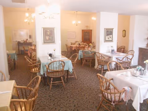 Dining room with several small tables covered in light tablecloths, wooden spindle-back chairs, and soft overhead lighting.