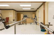 Physical therapy room with parallel bars for walking exercises, exercise equipment, a television, and medical posters on the walls in a healthcare facility.