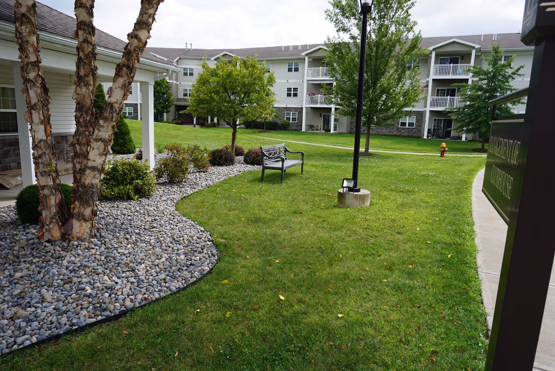 Outdoor view of a senior living facility with a well-maintained lawn, a bench, trees, and a multi-story residential building in the background. There is a pathway and a lamp post on the grass.