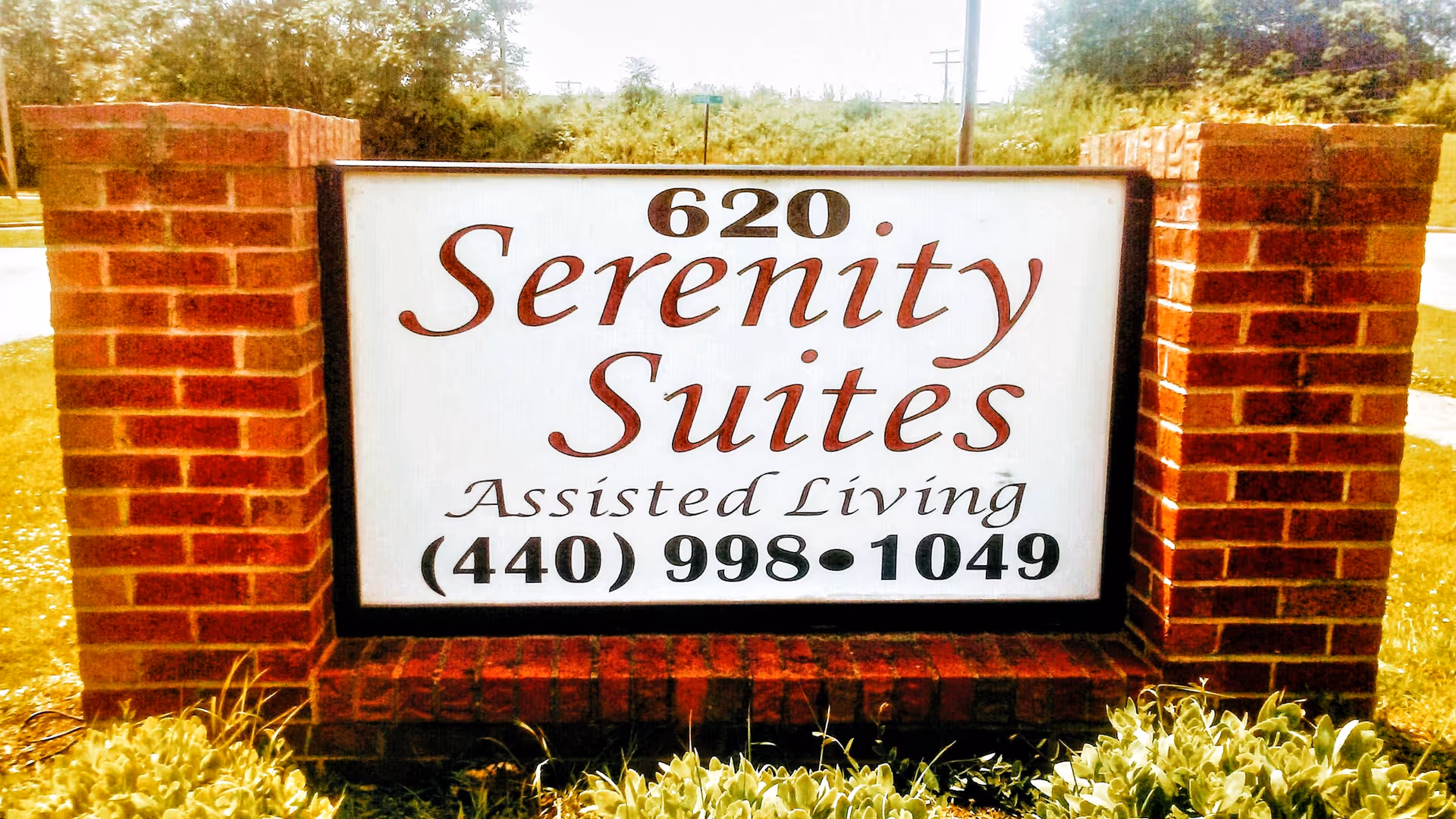 A brick signpost with a white rectangular sign displaying the text '620 Serenity Suites Assisted Living' and a phone number (440) 998-1049, situated outdoors with grass and trees in the background.