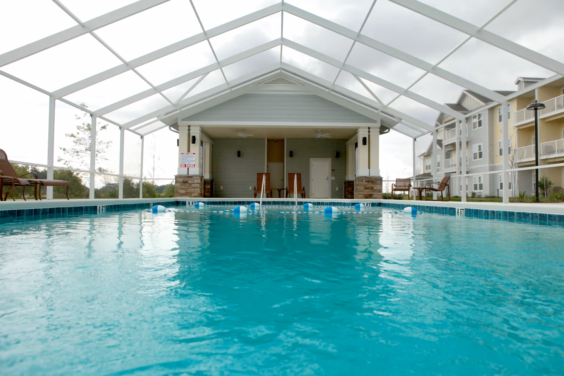 Indoor swimming pool with clear blue water under a white screened enclosure. There are lounge chairs and tables with chairs around the pool area. In the background, there is a small covered pavilion with ceiling fans and a door, and multi-story residential buildings are visible on the right side.