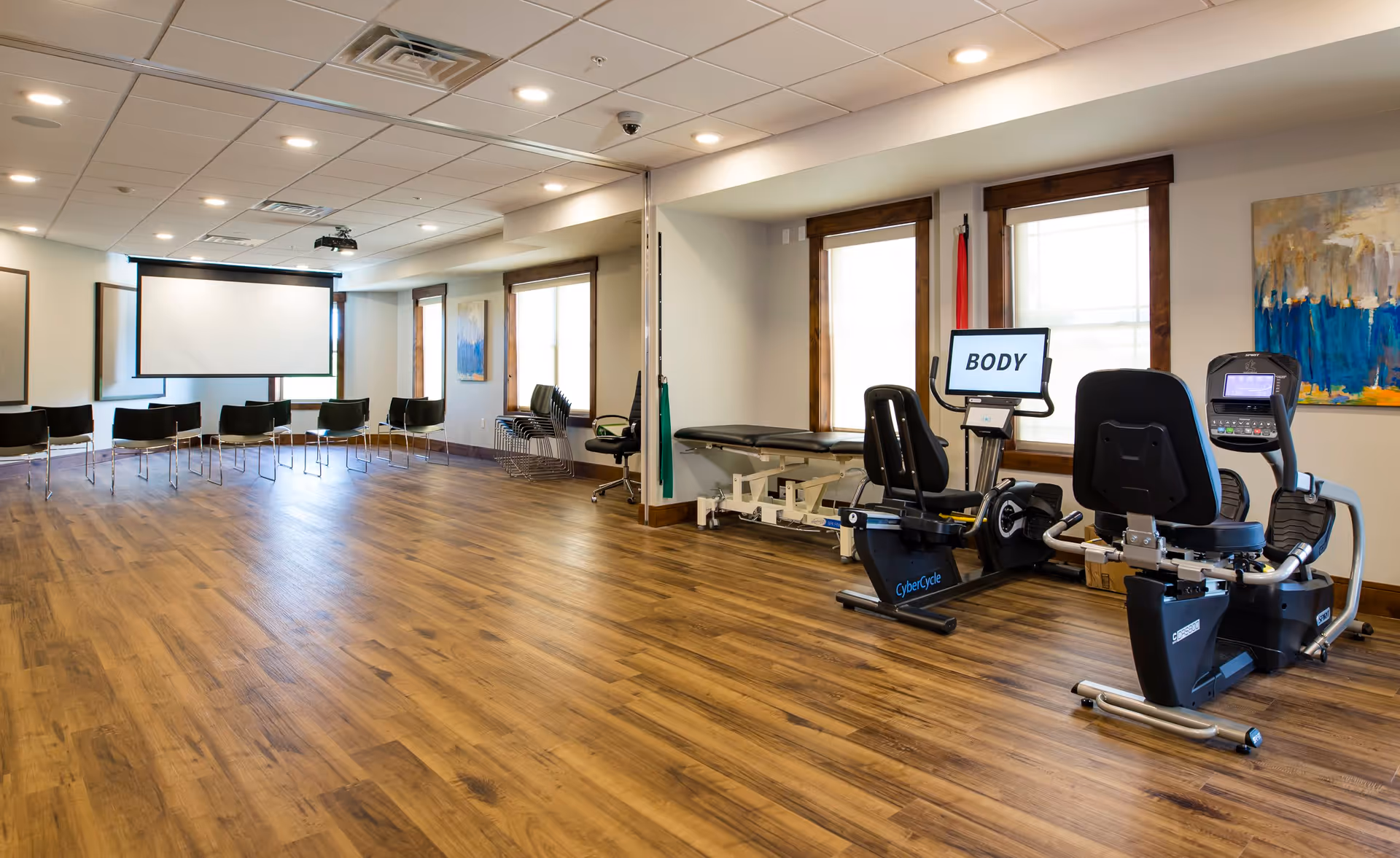 A spacious room with wooden flooring featuring exercise equipment including two recumbent bikes on the right side, a treatment table near the windows, and a row of chairs facing a large projector screen on the left side. The room is well-lit with ceiling lights and natural light from multiple windows. There is a colorful abstract painting on the wall and a monitor on one of the exercise bikes displaying the word 'BODY'.