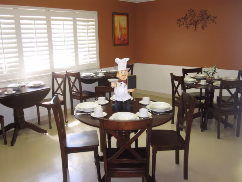 A dining room with several round wooden tables set with white plates, cups, and saucers. Each table has four wooden chairs. One table in the foreground has a decorative chef figurine holding a small chalkboard. The room has brown walls with a painting and a metal wall decoration, and white window shutters letting in natural light.