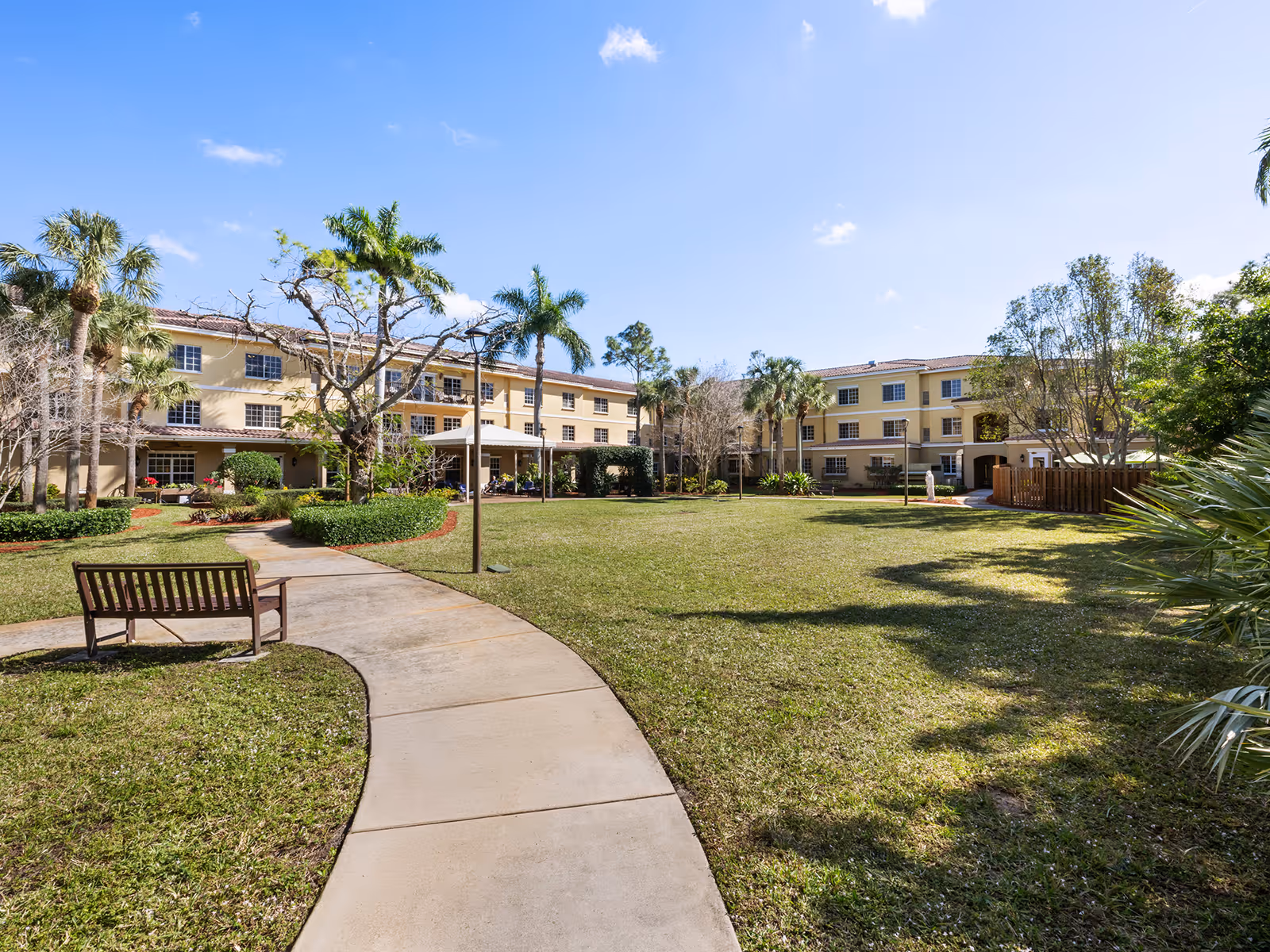 Outdoor view of Atria Park Of St. Joseph's senior living facility showing a large grassy courtyard with a curved concrete walkway, a wooden bench, palm trees, and a three-story beige building in the background under a clear blue sky.