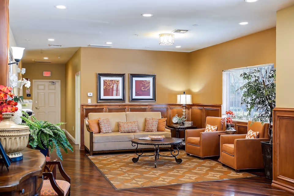 A cozy living room area with a beige sofa adorned with patterned cushions, two brown armchairs with chevron-patterned pillows, a round coffee table, and a side table with a lamp. The room has warm yellow walls, wooden wainscoting, framed artwork, a large window with blinds, and decorative plants.