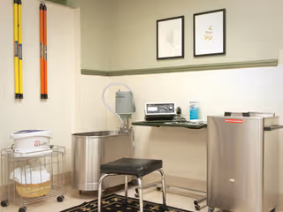 A clinical room with medical equipment including a stainless steel basin, a black padded stool, a small metal table with a radio and a bottle of hand sanitizer, and two framed botanical prints on the wall. There are also two long yellow and orange tools hanging on the wall and a white container with towels on a metal rack.
