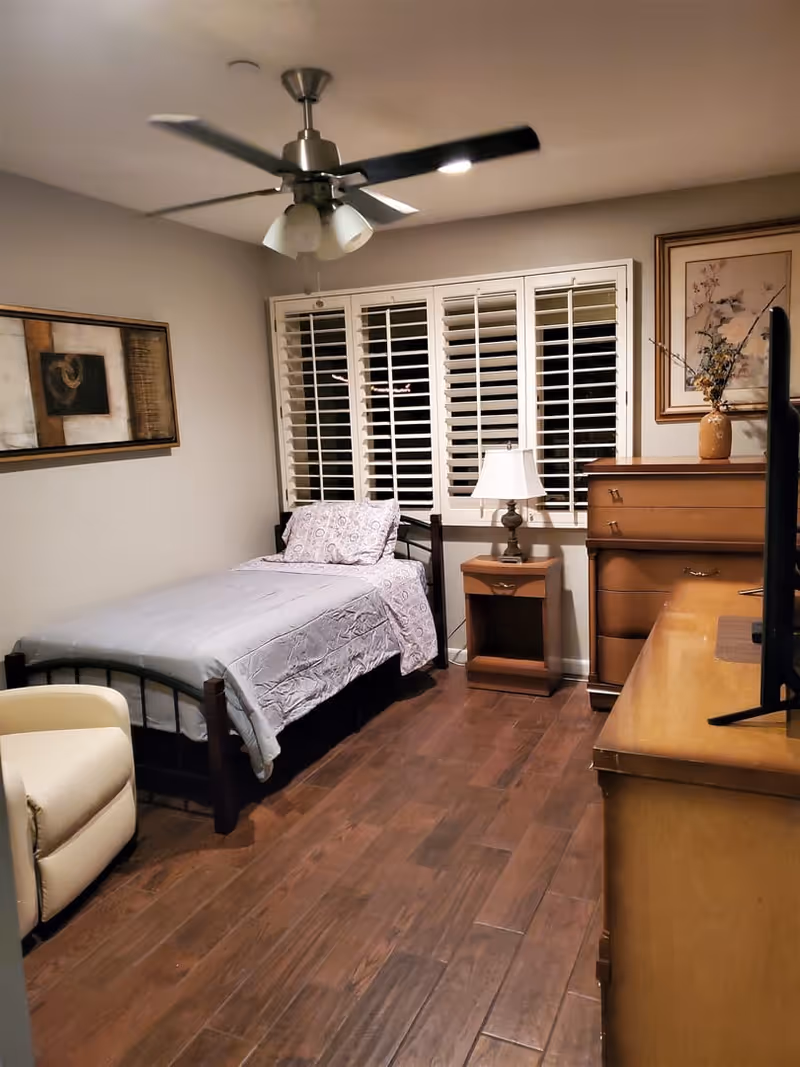 A cozy bedroom with a single bed covered in a light gray comforter and patterned pillow. There is a wooden nightstand with a lamp next to the bed, a wooden dresser with a vase and floral artwork above it, a ceiling fan with lights, and a cream-colored armchair. The room has wooden flooring and white plantation shutters on the window.