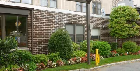 Brick exterior of a senior living center with manicured shrubs and flower beds along walkway.