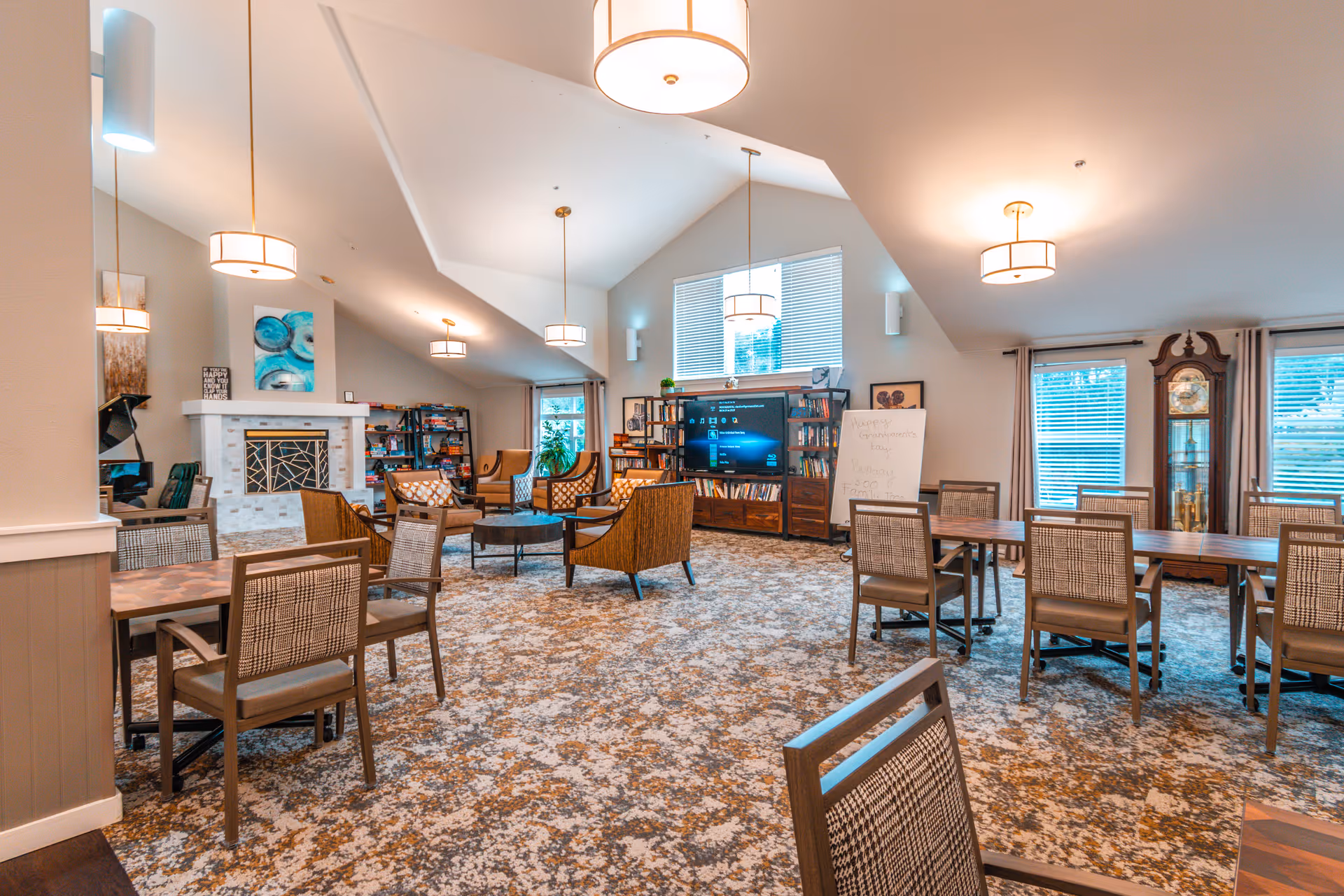Spacious, well-lit assisted living common room with tables and chairs, a seating area of armchairs around a TV and fireplace, bookshelves, and large windows.