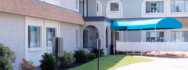 Exterior view of Northwood Rehabilitation & Health Care Center showing a beige stucco building with multiple windows, a blue awning over an entrance, a white picket fence, and a landscaped area with grass and bushes.