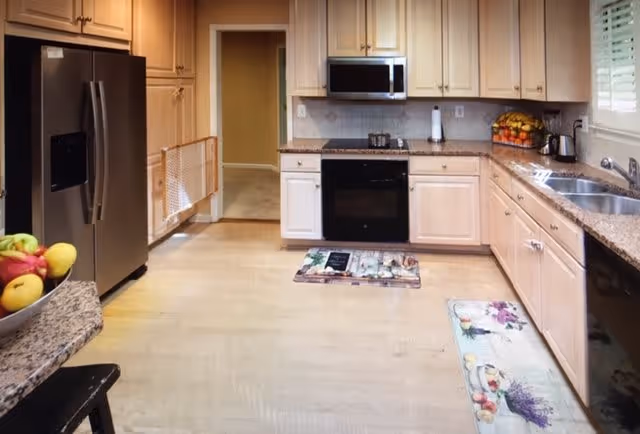 A spacious kitchen with light wood cabinets, granite countertops, and stainless steel appliances including a refrigerator, oven, and microwave. There are two decorative floor mats, a bowl of fruit on the counter, and a window with blinds above the sink.