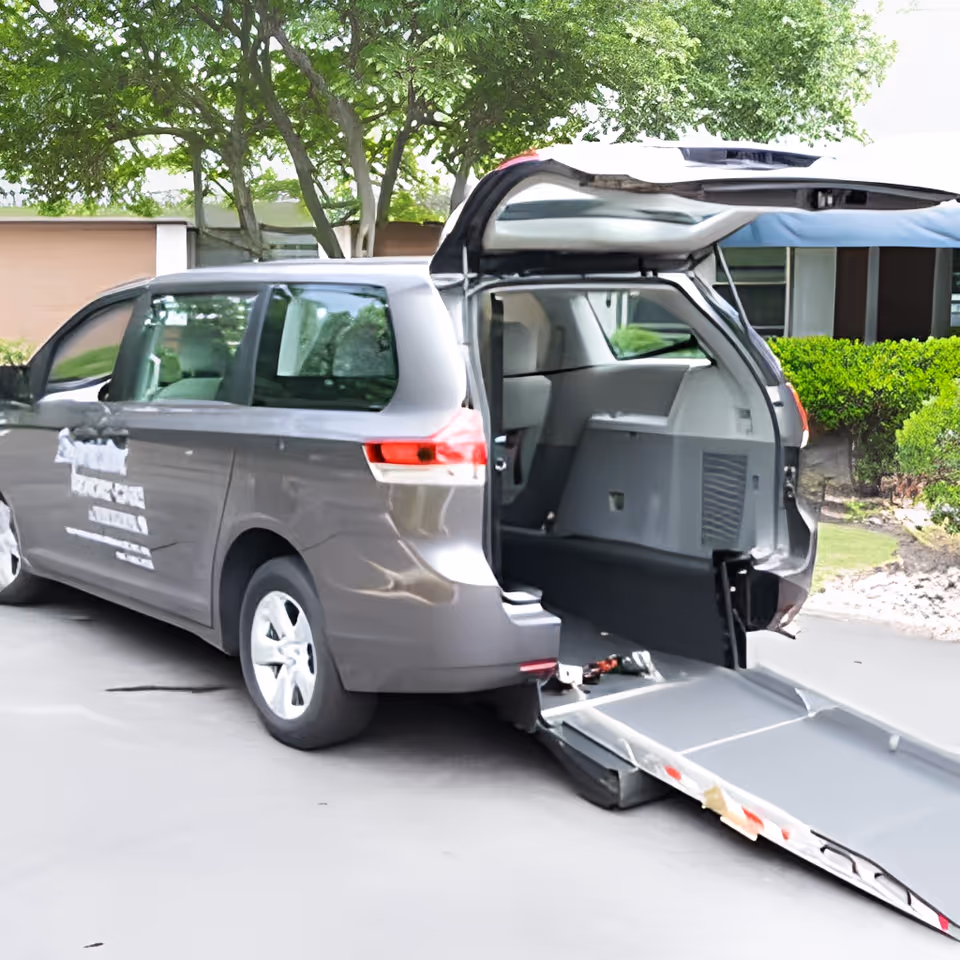 A gray wheelchair-accessible van with its rear hatch open and a ramp extended to the ground, parked outside near a building with greenery and trees in the background.