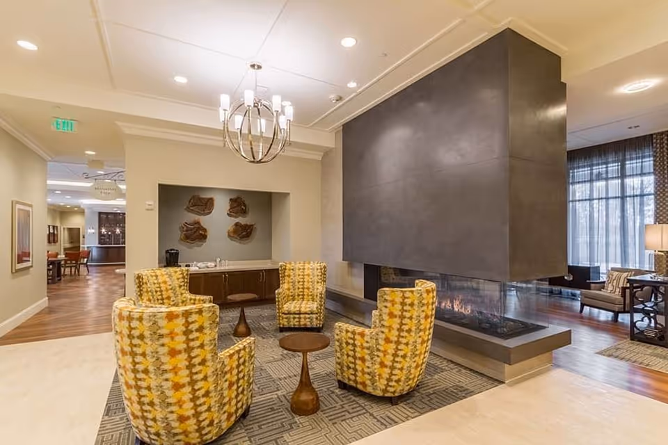 A cozy seating area in a senior living facility with four patterned yellow and orange armchairs arranged around two small wooden tables. A modern double-sided fireplace with a dark gray surround is the focal point, separating this area from another lounge space with additional seating and large windows with sheer curtains. The room features warm wood flooring, a chandelier, and soft lighting creating a welcoming atmosphere.