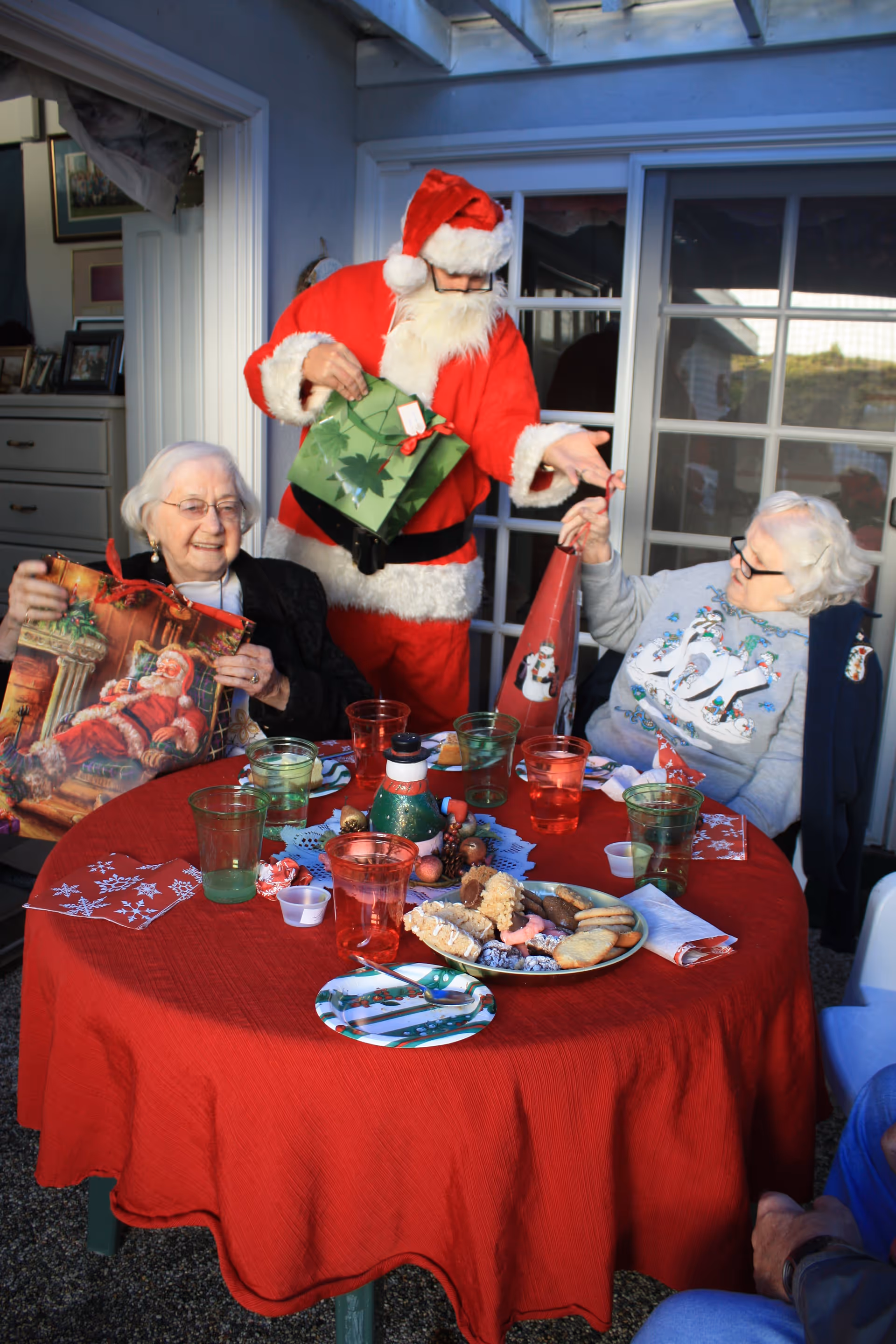 Two elderly women sitting at a round table covered with a red tablecloth, decorated with Christmas-themed napkins and plates with cookies. A person dressed as Santa Claus is standing behind them, handing a gift bag to one of the women. The setting appears to be a covered outdoor patio with glass doors in the background.