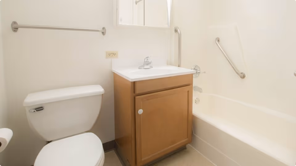 A small bathroom with a white toilet, a wooden vanity with a white sink and faucet, a bathtub with grab bars, and a towel bar on the wall.