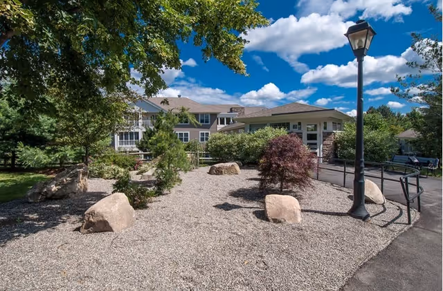 Outdoor garden area at Brookhaven At Lexington featuring a gravel landscape with large rocks, small trees and shrubs, a black lamp post, and a paved pathway leading to a building under a blue sky with scattered clouds.