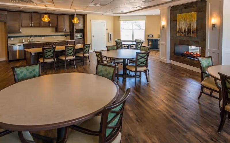 Communal dining room with round tables and chairs, a serving kitchen with barstools, and a wall fireplace.
