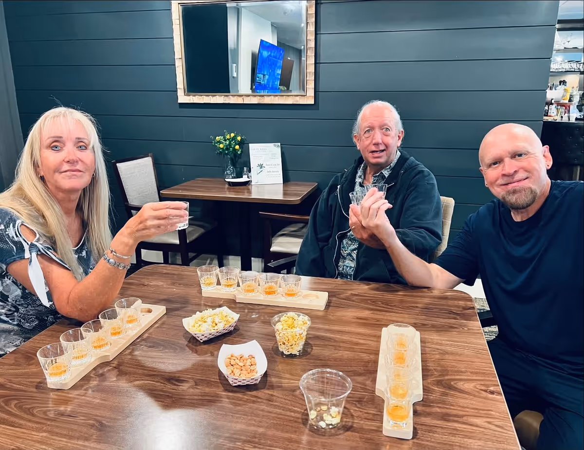 Three adults sitting around a wooden table in a room with dark paneled walls. They are holding small glasses, possibly for a tasting event. On the table are trays with several small glasses containing a light amber liquid, along with bowls of popcorn and nuts. A mirror and a TV are visible on the wall behind them.