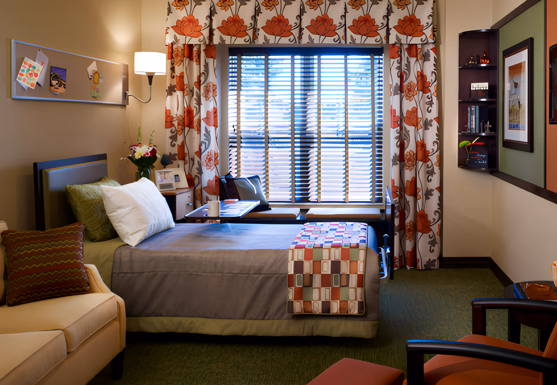 A cozy bedroom in a senior living facility with a single bed covered in gray bedding and a colorful patchwork quilt at the foot. The room features a large window with floral curtains and blinds, a beige sofa with a patterned pillow, a small side table with flowers and framed photos, a wall-mounted lamp, and a chair with an ottoman. The walls are decorated with a bulletin board and framed artwork.