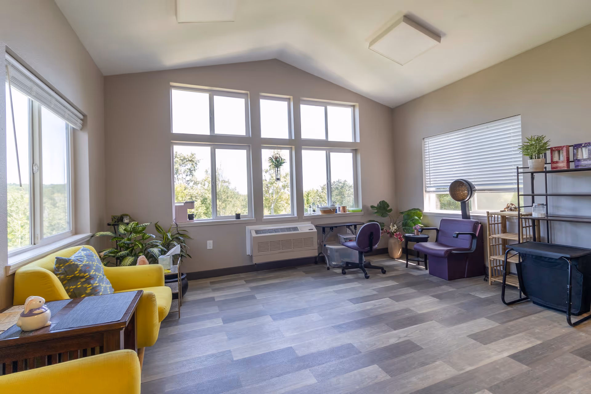 A bright room with large windows letting in natural light, featuring a yellow armchair with a patterned cushion, a small wooden side table, several potted plants, a desk with a purple office chair, a purple armchair with a floor lamp beside it, and a black metal shelving unit with various items on it. The room has light-colored walls and wood-patterned flooring.