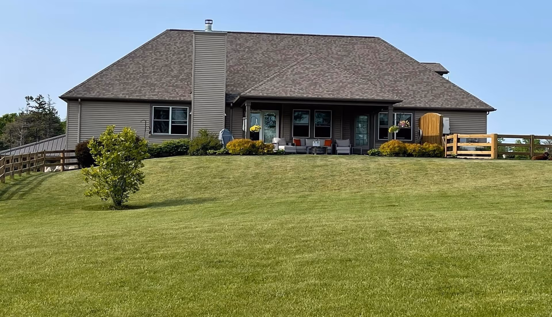 Single-story building with a covered front porch, outdoor seating, wooden fencing, and a wide green lawn in front.