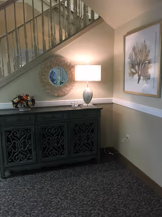 A corner of an interior space featuring a decorative dark green cabinet with intricate designs, a table lamp with a white shade, a round mirror with a textured frame hanging above the cabinet, a floral arrangement on the cabinet, and a framed artwork on the adjacent wall. A staircase with white railings is visible above the cabinet.