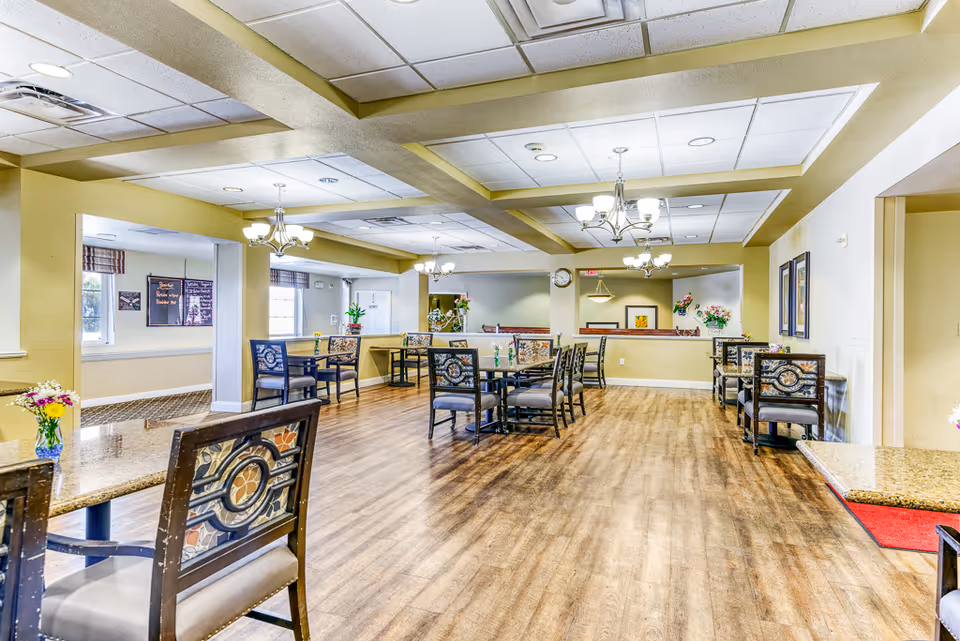 A spacious senior living dining room with wooden floors, multiple tables and chairs with decorative backs, chandeliers hanging from the ceiling, and floral arrangements on the tables. The room has light-colored walls and large windows allowing natural light to enter.