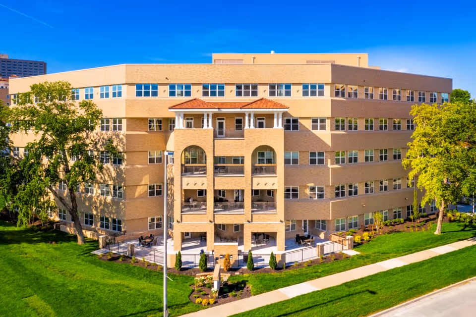 Exterior view of a large, multi-story senior living facility building with beige brick walls, numerous windows, and a central section featuring balconies and a red-tiled roof. The building is surrounded by green lawns, trees, and a sidewalk under a clear blue sky.