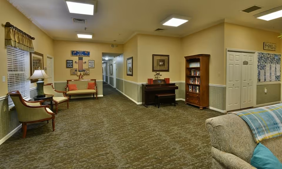 A cozy common area in an assisted living facility featuring a seating arrangement with chairs and a small sofa, a piano with a bench, a bookshelf filled with books, and framed pictures on the walls. The room has carpeted flooring, beige walls with a chair rail, and ceiling lights. A hallway extends from the room, and there are windows with curtains on the left side.