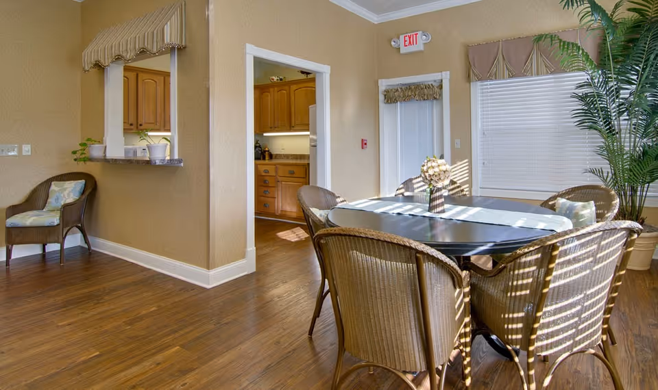A bright dining area in a senior living facility with a dark oval table surrounded by six wicker chairs. A vase with flowers is placed on the table. The room has wooden flooring, beige walls, a large window with blinds and a striped valance, and a tall potted plant in the corner. There is an open doorway leading to a kitchen with wooden cabinets, and a small pass-through window with two potted plants on the ledge. An exit sign is visible above a door in the background.