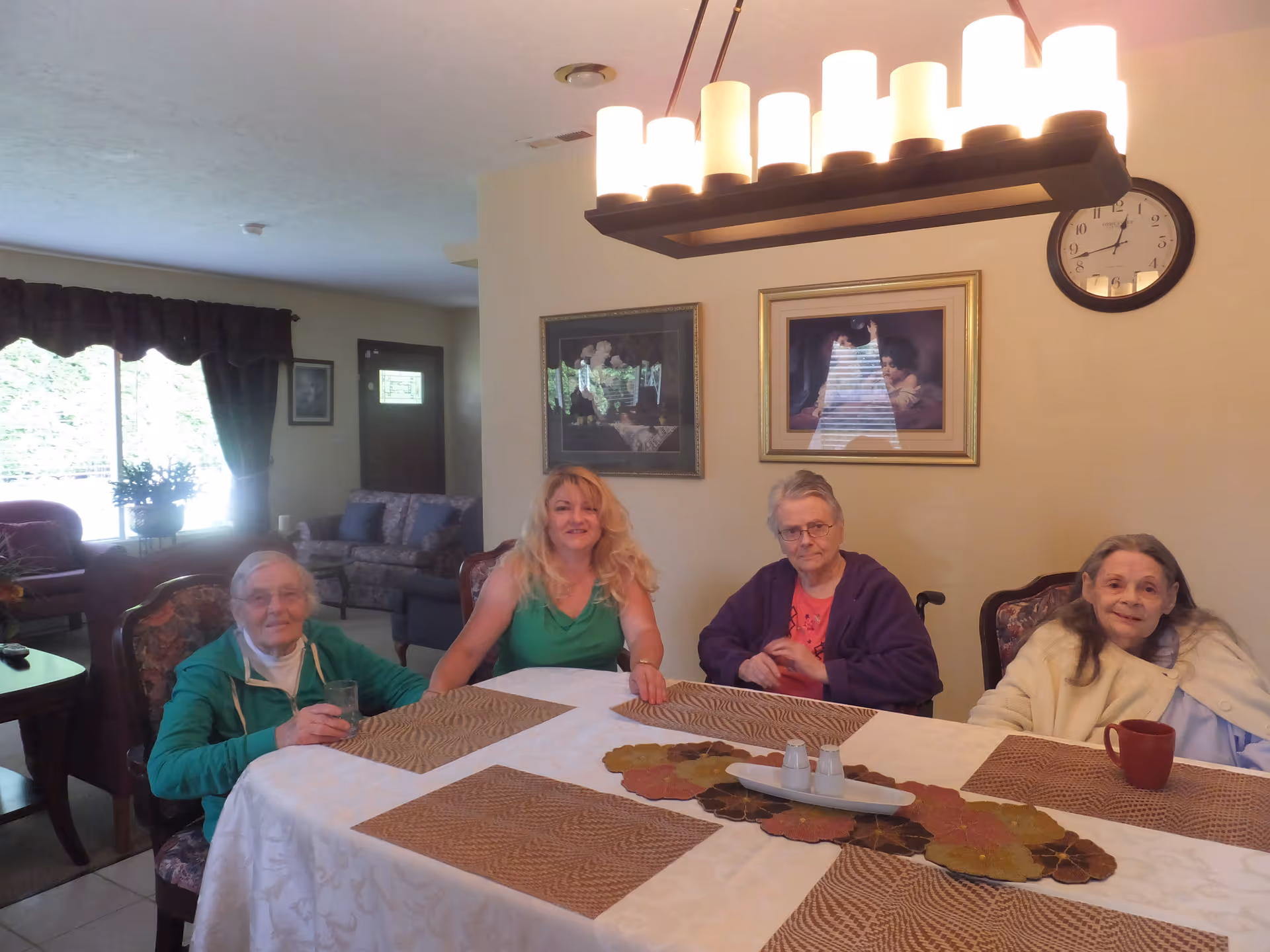 Four women sitting around a dining table in a well-lit room. The table is covered with a white tablecloth and brown placemats, with a decorative centerpiece and salt and pepper shakers. Behind them, there are framed pictures on the wall and a clock showing the time as 12:10. The room has a cozy living area with a window, curtains, and furniture visible in the background.