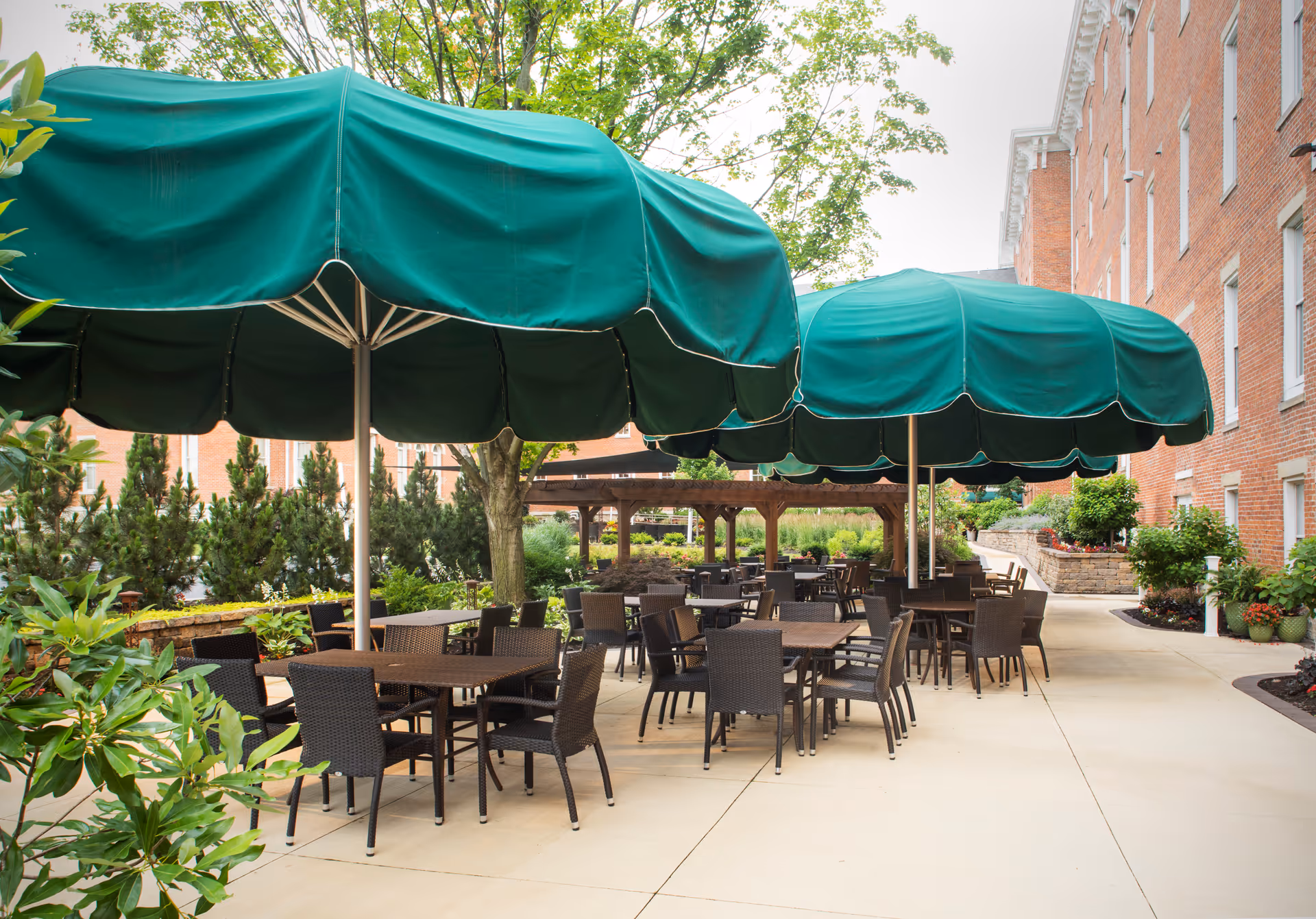Outdoor patio area at 10 Wilmington Place Retirement Community with multiple tables and chairs under large green umbrellas, surrounded by greenery and a brick building.