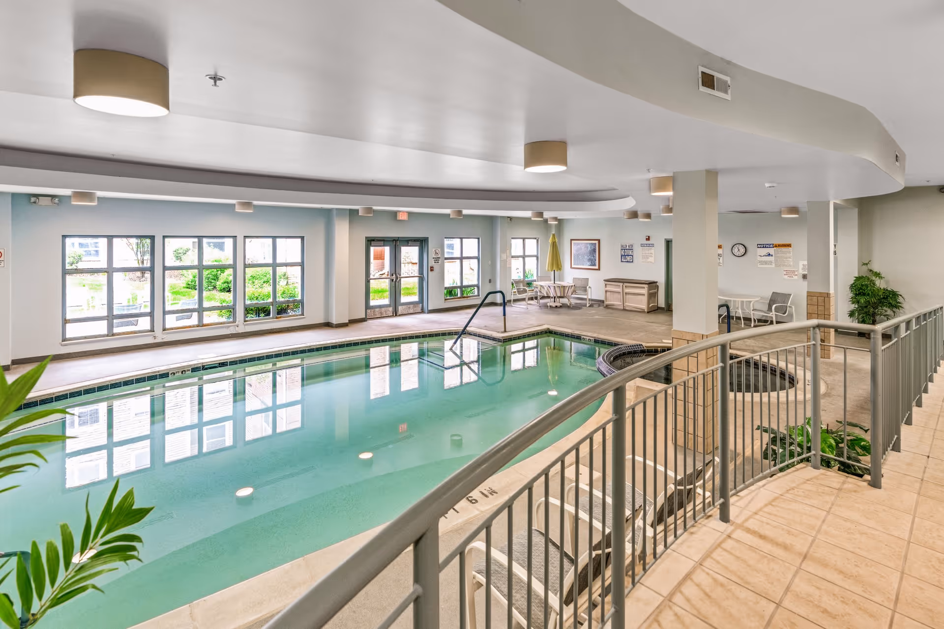 Indoor swimming pool area with clear water, surrounded by a tiled floor and metal railing. There are lounge chairs, tables with chairs, and potted plants around the pool. Large windows and glass doors provide natural light and a view of the outdoor greenery.