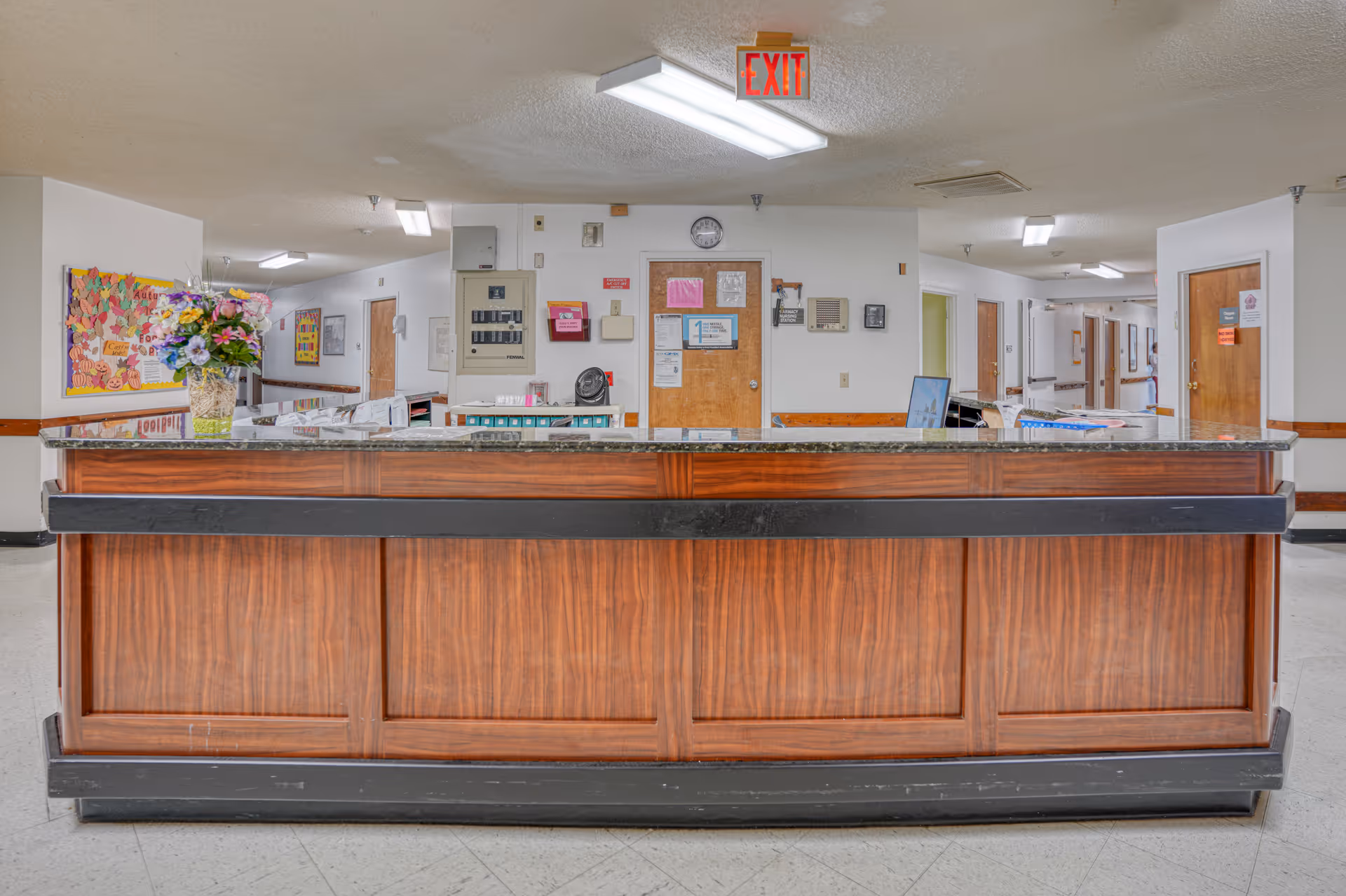 Reception desk area in a senior living facility hallway with a wooden counter, a vase of flowers on the left side, bulletin boards on the walls, and several closed doors along the corridor.