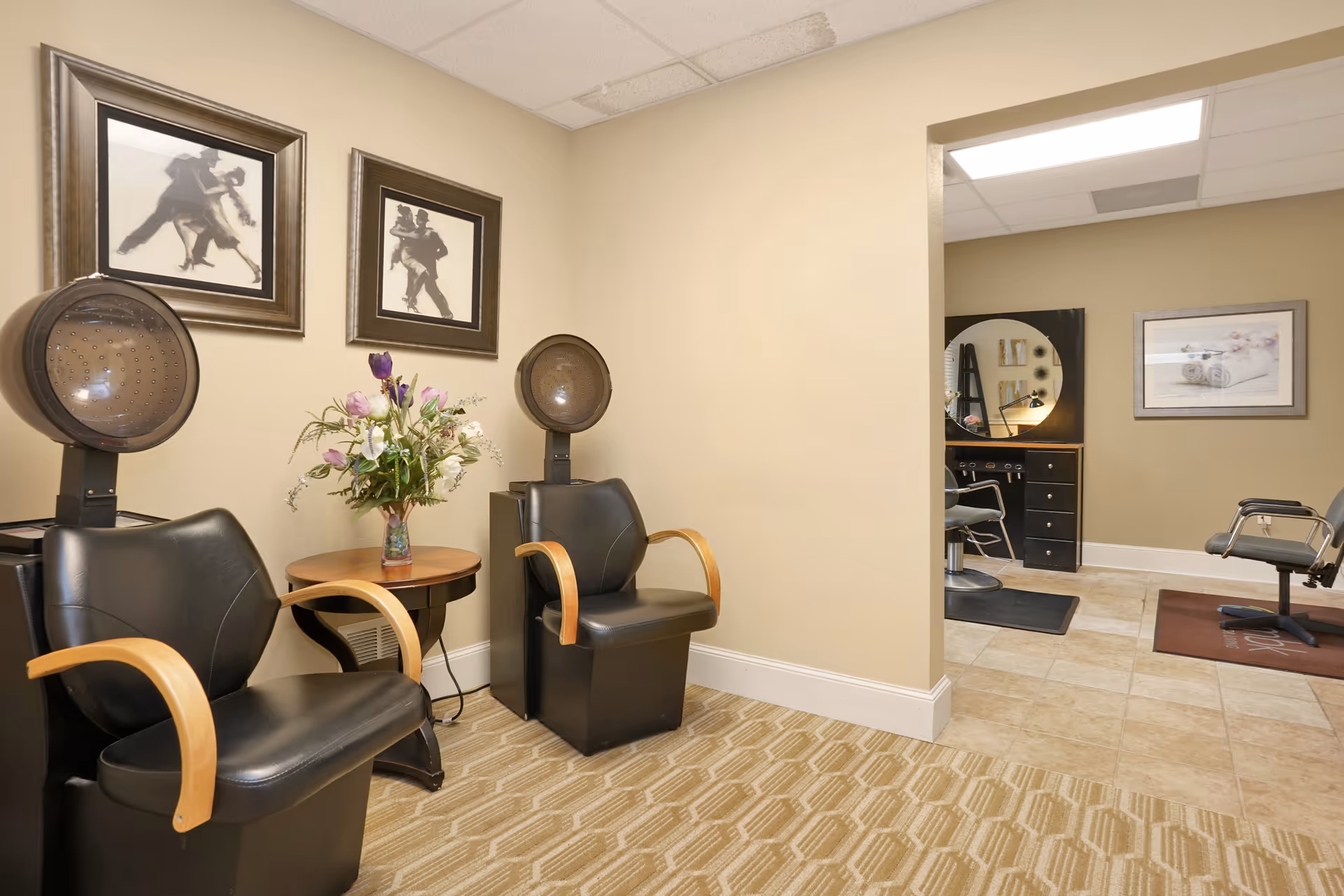 Interior view of a salon area in a senior living facility with two black salon chairs with wooden armrests and hair dryers above them. A small round table with a vase of flowers is placed between the chairs. The walls are beige with two framed pictures of dancing couples. In the background, there is another salon station with a round mirror, a chair, and a framed picture on the wall.