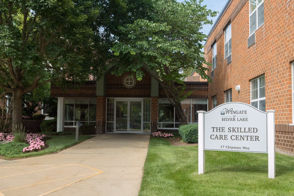 Entrance to Wingate at Silver Lake Skilled Care Center with a brick building, glass double doors, trees, and a sign displaying the facility name and address.
