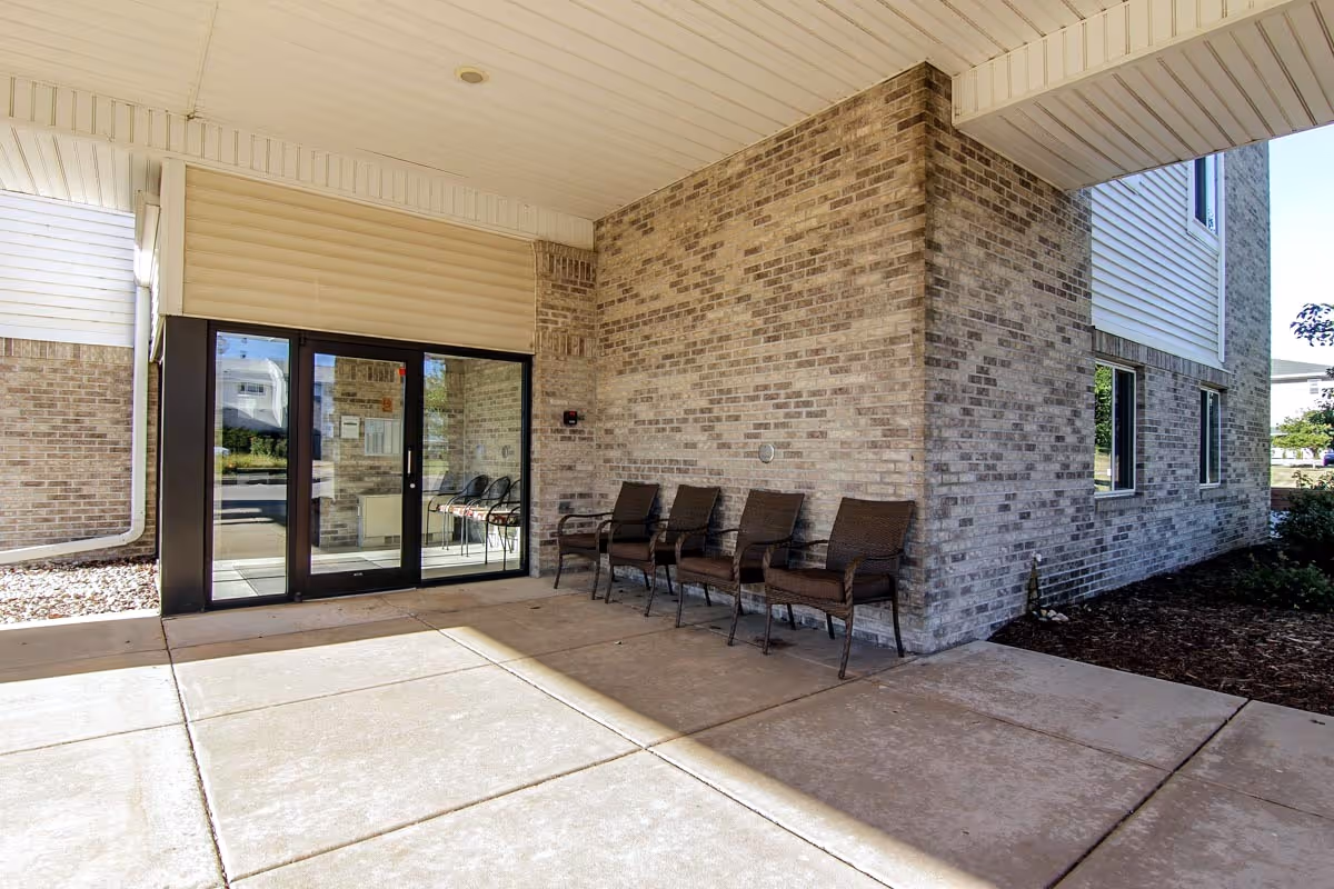 Covered entrance of a brick senior apartment building with glass double doors and a row of chairs along the wall.