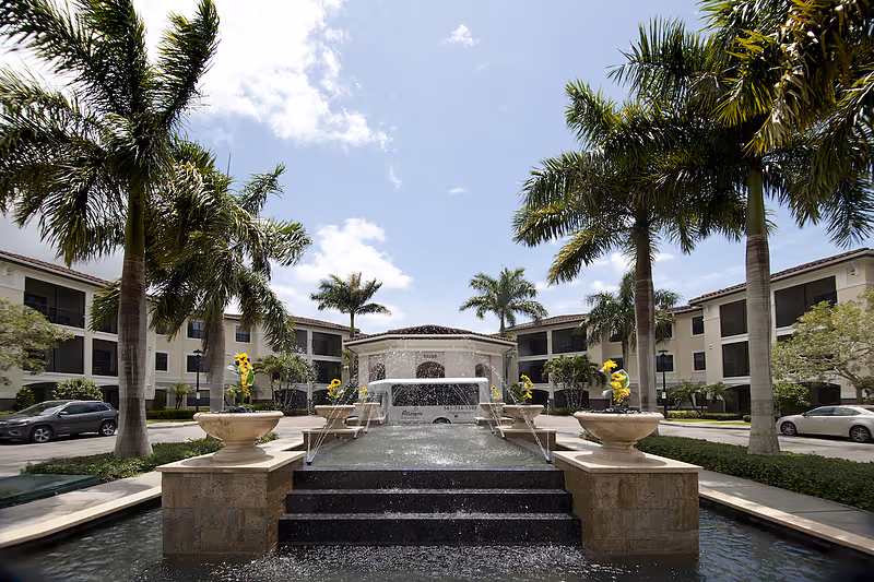 Exterior view of Allegro Senior Living in Boynton Beach, FL, featuring a central water fountain with water jets, surrounded by palm trees and a three-story building with balconies under a partly cloudy sky.