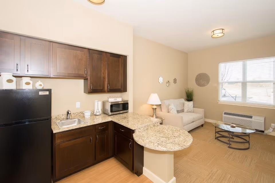 Interior view of a small living space in an assisted living facility showing a kitchenette with dark wood cabinets, a black refrigerator, a small sink, a microwave, and granite countertops extending into a small breakfast bar. Adjacent to the kitchenette is a cozy sitting area with a beige loveseat, a glass coffee table, a table lamp on a side table, decorative wall hangings, and a window with blinds letting in natural light.